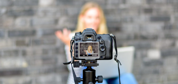Young woman in focus on digital camera LCD screen waving hand while seated on ground against wall for concept about video blogging